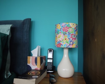 May include: A bedside table with a white lamp with a floral patterned lampshade. The lamp is on a wooden table with a book, a phone, a tissue box, and a pair of glasses. The wall behind the table is a bright turquoise blue.