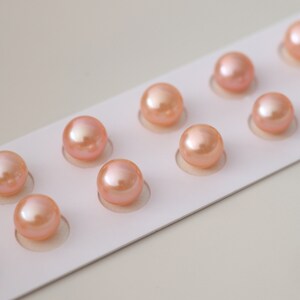May include: A close-up shot of a white card with multiple peach-colored pearls arranged in a row. The pearls have a lustrous sheen and are evenly spaced within individual circular cutouts. The background is a soft, neutral tone.