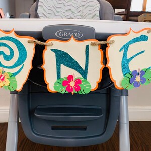May include: A 'ONE' banner with teal glitter letters on cream-coloured, scalloped cards with orange borders. The banner features paper flowers and tropical leaves, strung on twine. Ideal for a first birthday celebration, it is a colourful and festive decoration.
