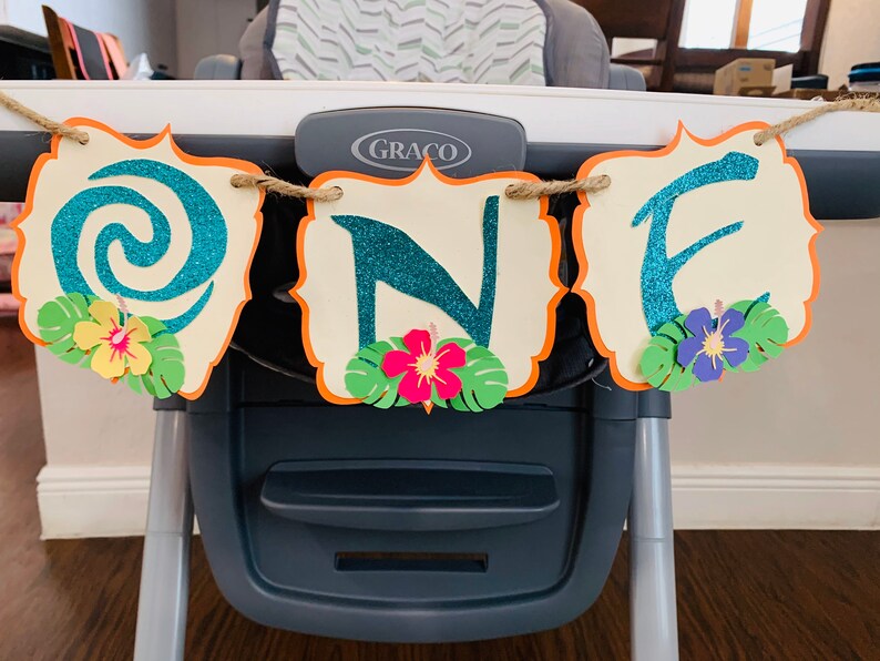 May include: A 'ONE' banner with teal glitter letters on cream-coloured, scalloped cards with orange borders. The banner features paper flowers and tropical leaves, strung on twine. Ideal for a first birthday celebration, it is a colourful and festive decoration.