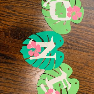 May include: Three decorative paper cutouts spelling "LIFE". Each letter is white and set on a green monstera leaf with a pink hibiscus flower. The cutouts are arranged on a brown wooden surface.