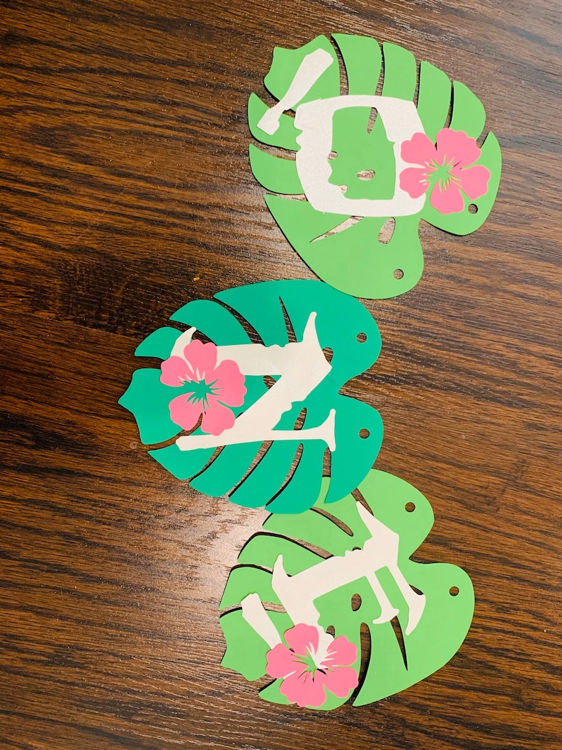 May include: Three decorative paper cutouts spelling "LIFE". Each letter is white and set on a green monstera leaf with a pink hibiscus flower. The cutouts are arranged on a brown wooden surface.
