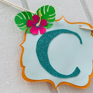 May include: A decorative banner featuring a teal glitter letter 'C' on a light blue background. The banner is adorned with a pink hibiscus flower and green leaves. It is strung on a white string.