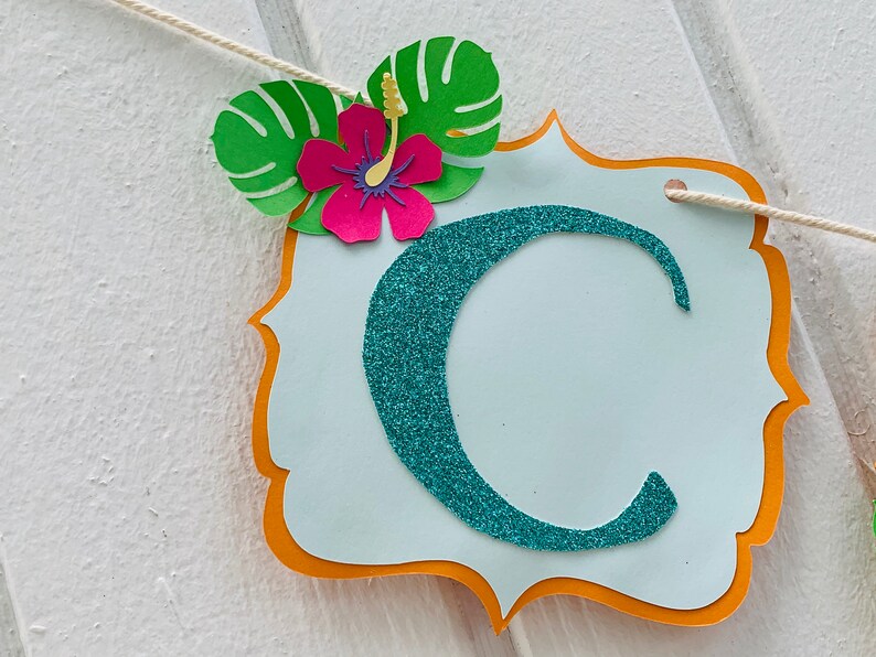 May include: A decorative banner featuring a teal glitter letter 'C' on a light blue background. The banner is adorned with a pink hibiscus flower and green leaves. It is strung on a white string.