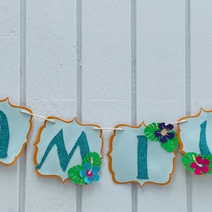 May include: A banner spelling "CAMILA" in teal glitter letters on light blue cutouts with gold trim. Each cutout is decorated with tropical flowers and leaves, strung on a white string. Ideal for a birthday party.