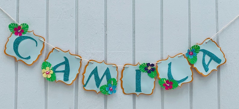 May include: A banner spelling "CAMILA" in teal glitter letters on light blue cutouts with gold trim. Each cutout is decorated with tropical flowers and leaves, strung on a white string. Ideal for a birthday party.