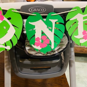 May include: A decorative banner with green leaf cutouts, white letters, and pink flower accents. The banner spells out the word "ONE" and is strung on a natural twine. The banner is hanging in front of a high chair.