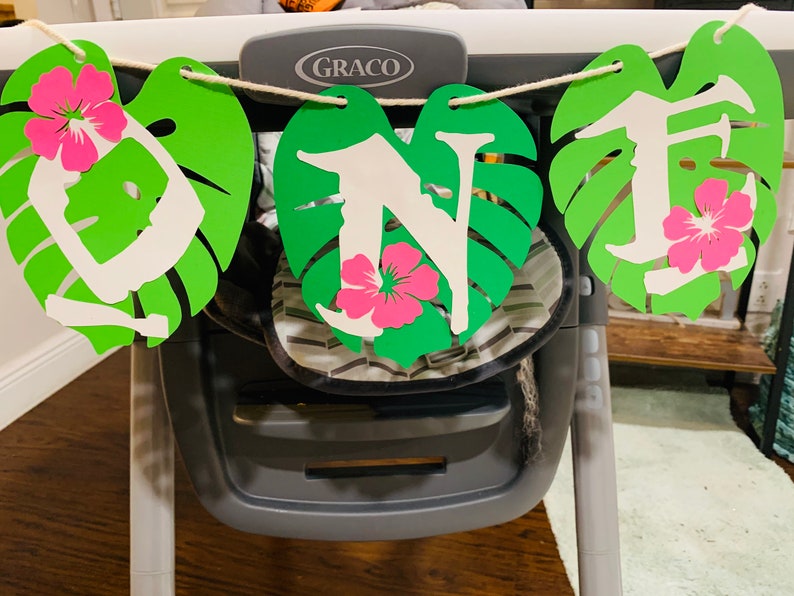 May include: A decorative banner with green leaf cutouts, white letters, and pink flower accents. The banner spells out the word "ONE" and is strung on a natural twine. The banner is hanging in front of a high chair.