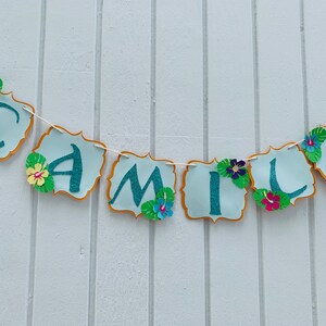 May include: A banner spelling "CAMILA" in teal glitter letters on light blue scalloped shapes with gold trim. The banner is decorated with paper flowers and tropical leaves, strung on a white string.
