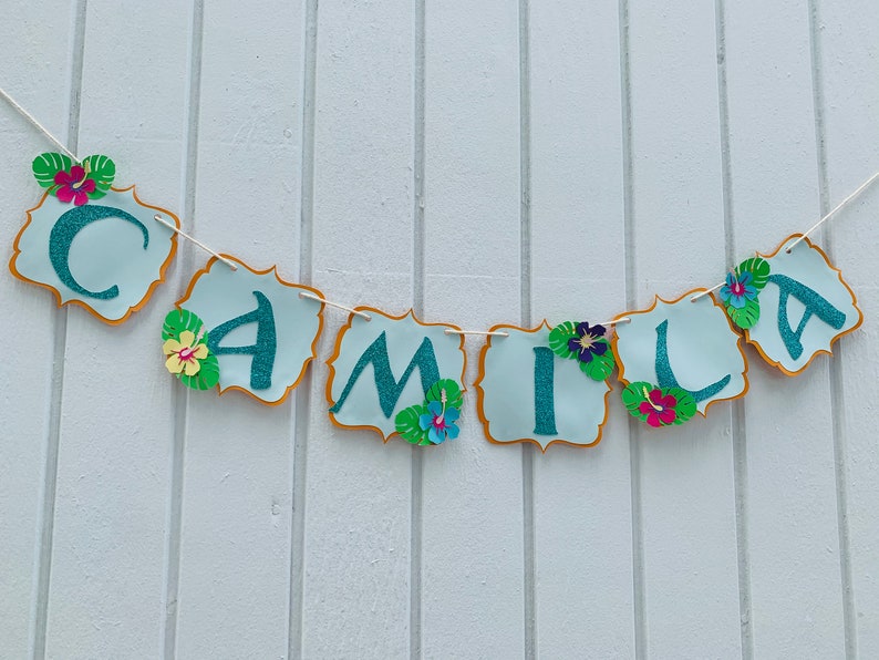 May include: A banner spelling "CAMILA" in teal glitter letters on light blue scalloped shapes with gold trim. The banner is decorated with paper flowers and tropical leaves, strung on a white string.