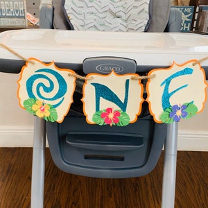 May include: A decorative banner spelling "ONE" in teal glitter letters, with a tropical theme. Each letter is on a cream-coloured, scalloped-edge shape with an orange border. The banner is adorned with paper flowers and leaves, strung on twine.