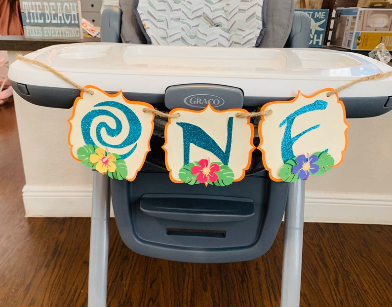 May include: A decorative banner spelling "ONE" in teal glitter letters, with a tropical theme. Each letter is on a cream-coloured, scalloped-edge shape with an orange border. The banner is adorned with paper flowers and leaves, strung on twine.
