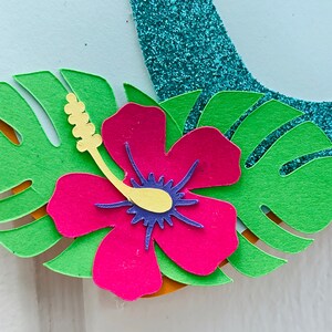 May include: Close-up of a tropical paper craft design. It features a vibrant pink hibiscus flower with a yellow stamen and blue centre, surrounded by green monstera leaves. A turquoise glitter element is visible.