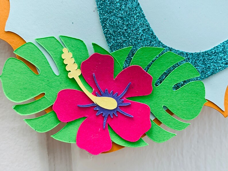 May include: Close-up of a tropical paper craft design. It features a vibrant pink hibiscus flower with a yellow stamen and blue centre, surrounded by green monstera leaves. A turquoise glitter element is visible.