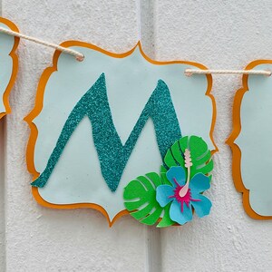 May include: A banner with a pale blue background and an orange border. The letter "M" is in the centre, made of teal glitter. A paper flower and green leaves are attached to the banner. The banner is strung on natural twine.