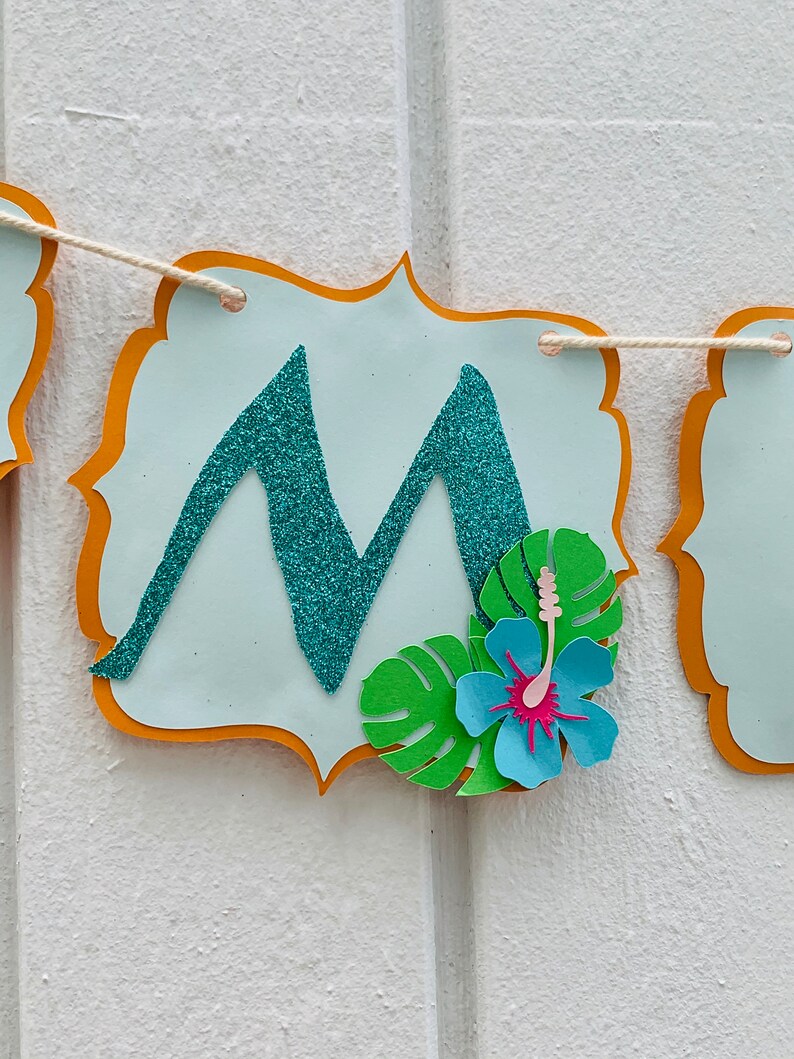 May include: A banner with a pale blue background and an orange border. The letter "M" is in the centre, made of teal glitter. A paper flower and green leaves are attached to the banner. The banner is strung on natural twine.