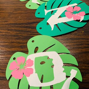 May include: Green paper cutouts in the shape of monstera leaves, each with a white silhouette and a pink hibiscus flower. The silhouettes appear to be letters. The leaves are arranged on a dark wood surface.