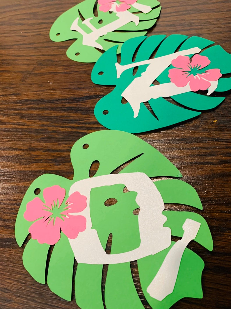 May include: Green paper cutouts in the shape of monstera leaves, each with a white silhouette and a pink hibiscus flower. The silhouettes appear to be letters. The leaves are arranged on a dark wood surface.