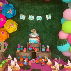 May include: A birthday party setup featuring a cake, cupcakes, and decorations. The cake has a blue and white ombre design with a topper that says "Camila". Cupcakes are arranged on wooden crates. Balloons and large paper flowers add to the festive atmosphere.