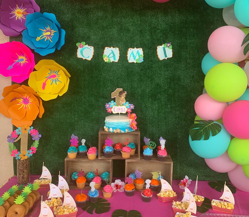 May include: A birthday party setup featuring a cake, cupcakes, and decorations. The cake has a blue and white ombre design with a topper that says "Camila". Cupcakes are arranged on wooden crates. Balloons and large paper flowers add to the festive atmosphere.