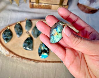 May include: A collection of labradorite gemstone pendants with gold-toned accents. The pendants are displayed on a wooden surface. The labradorite stones have a blue, green, and gold iridescent sheen.