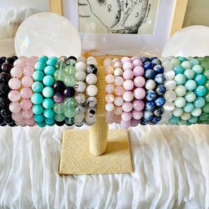 May include: A display of colourful beaded bracelets made with natural stones. The bracelets are arranged on a beige stand and feature various colours, including blue, green, pink, and purple.
