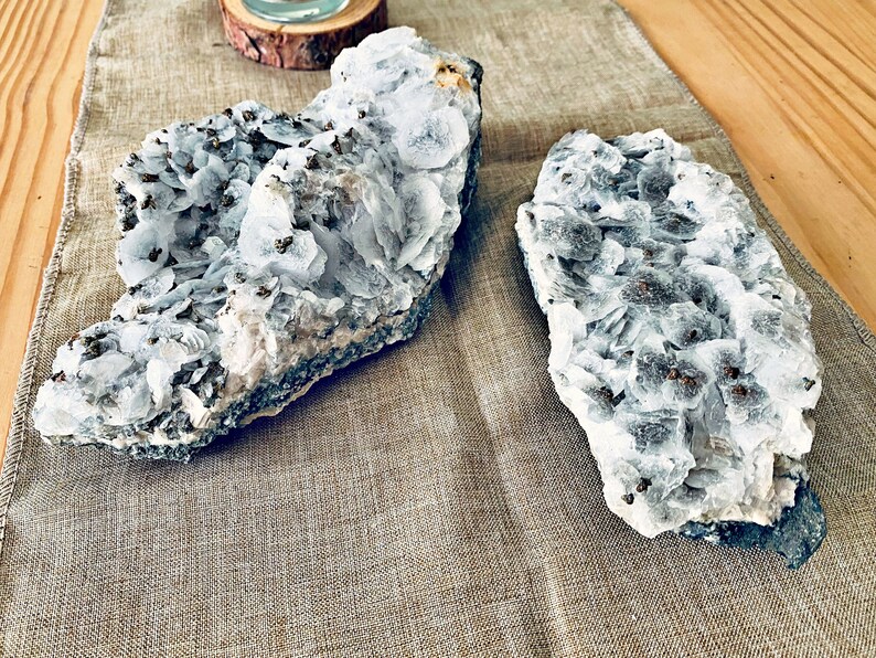 May include: Two pieces of white selenite crystal clusters with a gray, rough, natural base. The crystals are clustered together in a natural formation.