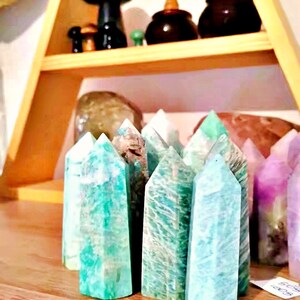 May include: A collection of green, blue, and purple crystal points on a wooden shelf. The crystals are arranged in a row and are all pointed upwards.