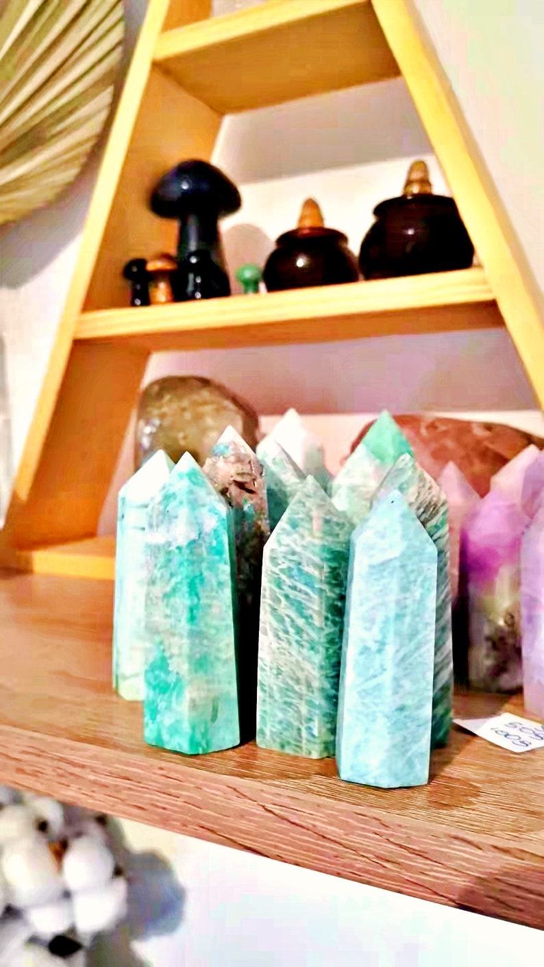 May include: A collection of green, blue, and purple crystal points on a wooden shelf. The crystals are arranged in a row and are all pointed upwards.