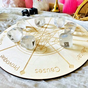 May include: A wooden circular board with a mandala design and eight points. Each point has a word written on it: "Chance", "Provoque", "Causes", "Aeviter", "Resultat",  "Chance", "Provoque", "Causes".  There are eight clear crystal dice with black symbols on them placed on the board.