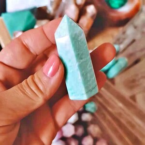 May include: A light green, pointed crystal with a smooth, polished surface. The crystal is held in a hand, with other crystals visible in the background.