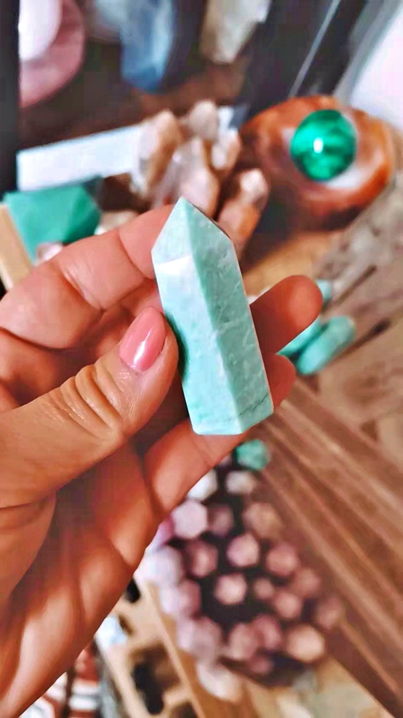 May include: A light green, pointed crystal with a smooth, polished surface. The crystal is held in a hand, with other crystals visible in the background.