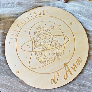 May include: A round wooden plaque with a laser-engraved design of a crystal cluster surrounded by a circle and stars. The text "LES CRISTAUX d'Ana" is engraved around the design.