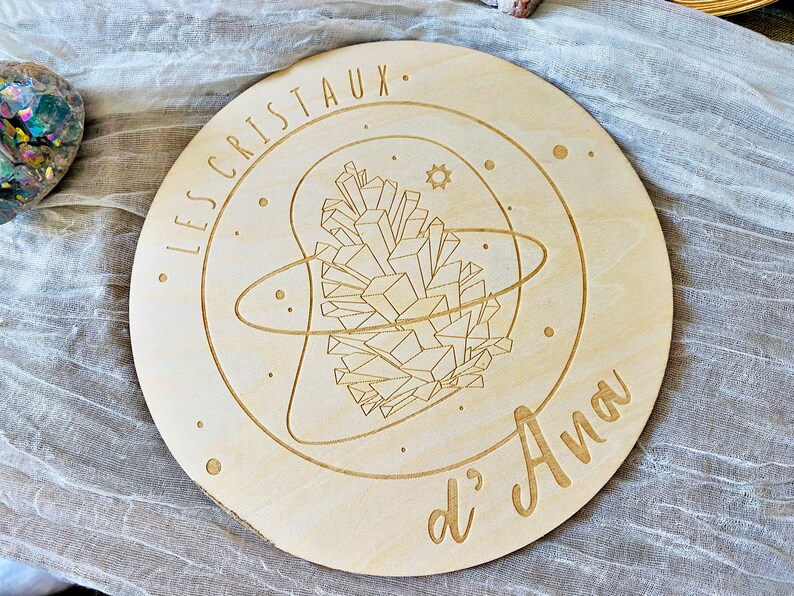 May include: A round wooden plaque with a laser-engraved design of a crystal cluster surrounded by a circle and stars. The text "LES CRISTAUX d'Ana" is engraved around the design.
