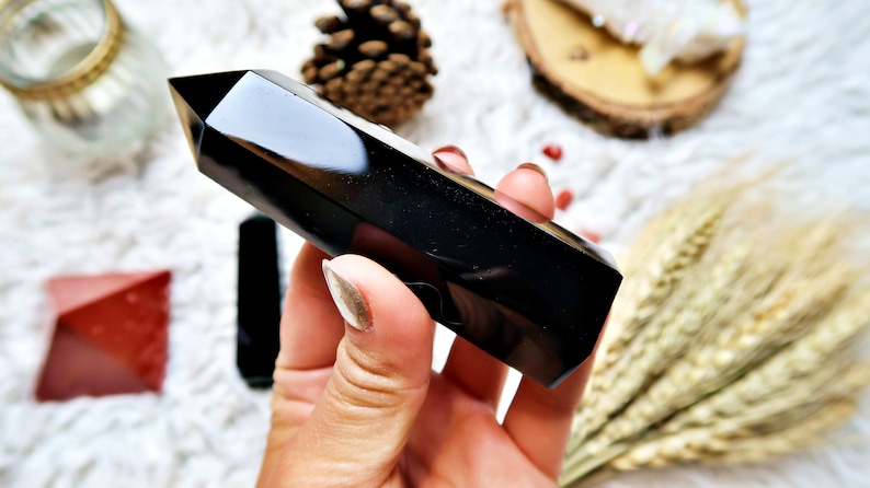 Pu&ograve; includere: Una punta di cristallo di ossidiana nera lucidata. Il cristallo &egrave; tenuto in mano e ha una superficie liscia e riflettente. Altri cristalli e oggetti decorativi sono sullo sfondo.