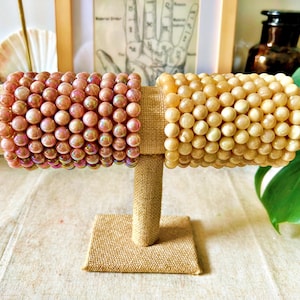 Peut inclure: Deux bracelets de perles exposés sur un présentoir en toile de jute. L'un est rose avec des perles irisées, l'autre est de couleur crème. Le présentoir est sur une surface claire, avec une image encadrée et de la verdure en arrière-plan.