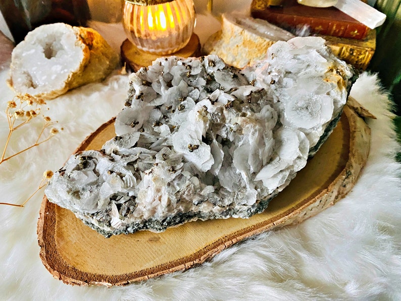 May include: A large, white, and gray crystal cluster with a rough, natural texture. The cluster is covered in small, gold-colored pyrite crystals. The cluster is sitting on a wooden slice.