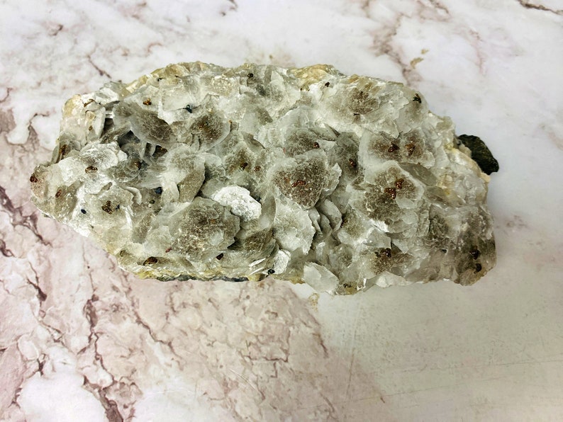 May include: A cluster of white and gray mica crystals with small, reddish-brown inclusions. The crystals are arranged in a rough, irregular shape.