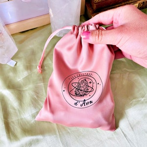 May include: A pink velvet drawstring pouch with a black logo that says "Les Cristaux d'Ana" and a stylized pine cone design.