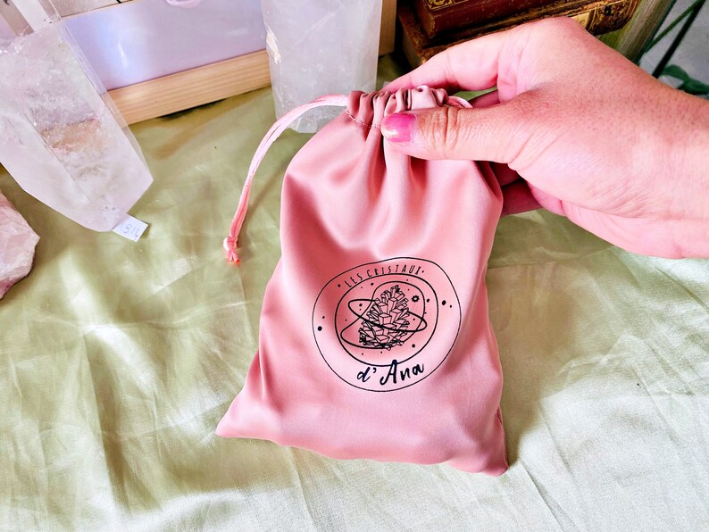 May include: A pink velvet drawstring pouch with a black logo that says "Les Cristaux d'Ana" and a stylized pine cone design.