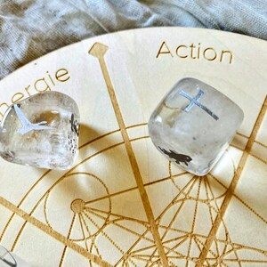 May include: Two clear quartz crystals with silver symbols on a wooden board with the words "Action" and "Energie" inscribed. The board has a geometric pattern.