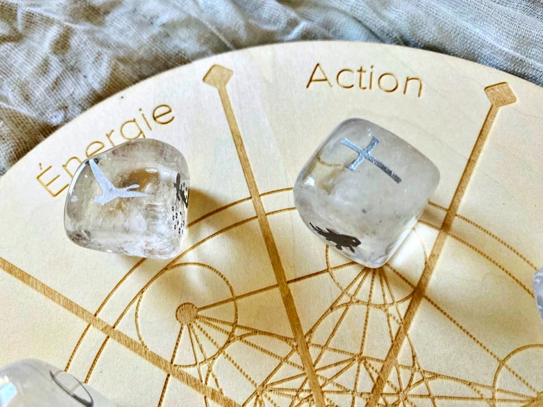 May include: Two clear quartz crystals with silver symbols on a wooden board with the words "Action" and "Energie" inscribed. The board has a geometric pattern.