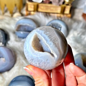 Peut inclure: Gros plan sur une sphère d'agate bleue avec une druse de cristal naturelle à l'intérieur. La sphère est lisse et polie, de couleur bleu clair et avec une druse blanche à l'intérieur. La sphère est tenue dans une main.