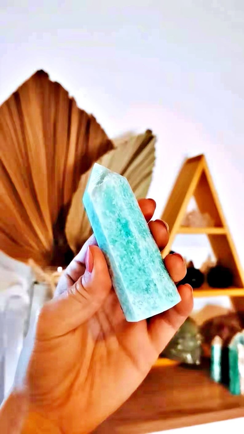 May include: A light blue, translucent, crystal point with a rough, textured surface. The crystal is held in a hand.