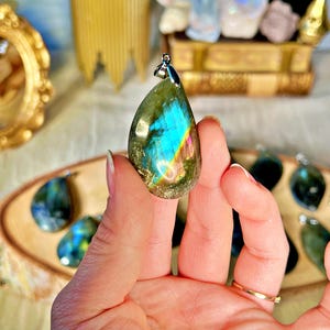 May include: A teardrop-shaped labradorite pendant with iridescent blue, green, and gold hues. The pendant is held in a hand and has a silver bail.