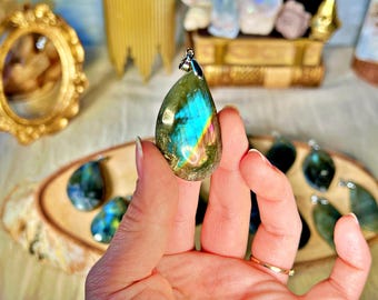May include: A teardrop-shaped labradorite pendant with iridescent blue, green, and gold hues. The pendant is held in a hand and has a silver bail.