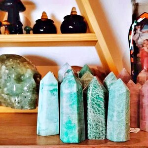 May include: A collection of green and white polished crystal points, also known as green aventurine, arranged on a wooden shelf.