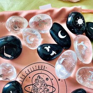 May include: A collection of smooth, polished stones with various etched designs, including a heart, a key, a crescent moon, a sun, a bird, and a crescent moon. The stones are in a pink pouch with the text "LES CRISTAUX" printed on it.