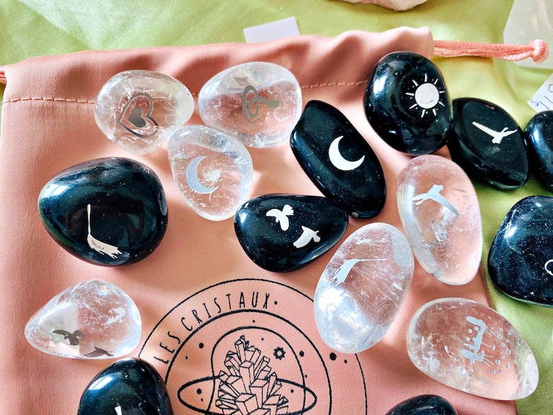 May include: A collection of smooth, polished stones with various etched designs, including a heart, a key, a crescent moon, a sun, a bird, and a crescent moon. The stones are in a pink pouch with the text "LES CRISTAUX" printed on it.