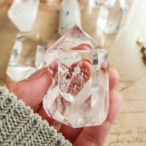 Peut inclure: Une pointe de cristal de quartz clair avec une double terminaison. Le cristal est tenu dans une main et est entouré d'autres cristaux.